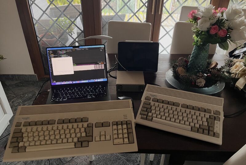Two Amiga computers on a table, with a laptop and other modern devices behind them.